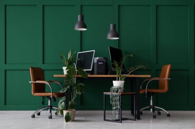 İş yerinde bilgisayar monitörleri, taşınabilir elektrik santrali ve ofiste yeşil duvarın yanında fabrikalar var.