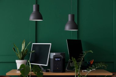 İş yerinde bilgisayar monitörleri, taşınabilir elektrik santrali ve ofiste yeşil duvarın yanında fabrikalar var.