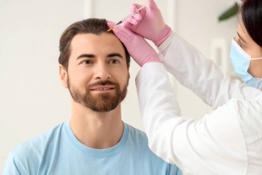 Genç adam trikoloğun ofisinde saç büyütme iğnesi yaptırıyor.