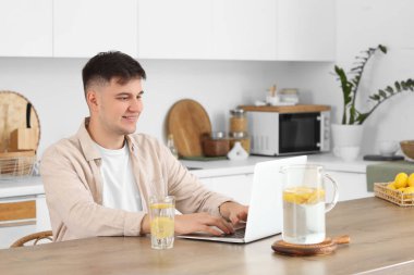 Mutfaktaki masada dizüstü bilgisayar kullanarak limonlu su içen genç adam.