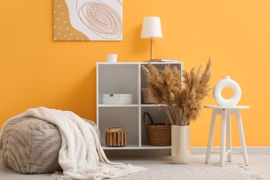 Ekoseli gri pouf, raf ünitesi ve vazoda sarı duvarın yanındaki vazoda otlar.