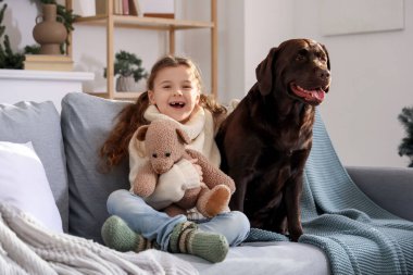 Labrador köpeği ve oyuncak ayısıyla evdeki kanepede oturan sevimli mutlu kız.