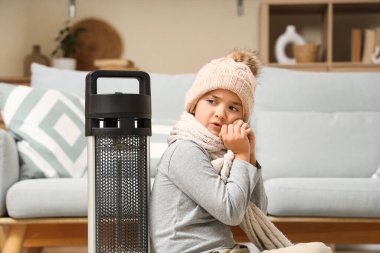 Frozen boy in warm hat with electric heater at home clipart