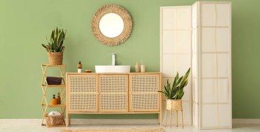 Seramik lavabo, ayna ve çekmeceler şık banyoda yeşil duvarın yanında.