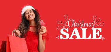 Noel Baba şapkalı güzel Afro-Amerikalı kadın elinde alışveriş çantaları ve kırmızı arka planda hediye kartı tutuyor. Noel indirimi