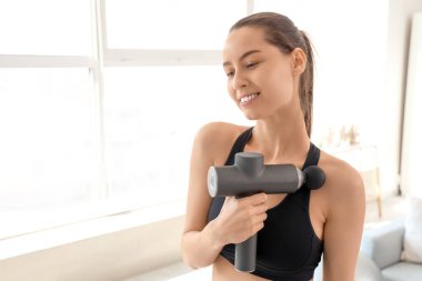 Sporty young woman massaging her shoulder with percussive massager at home