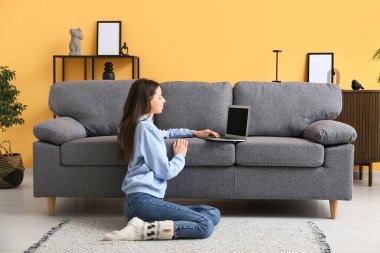 Genç bir kadın kanepenin yanında oturuyor ve oturma odasında dizüstü bilgisayar kullanıyor.