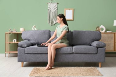 Genç ve güzel bir kadın kanepede oturuyor ve şık bir oturma odasında dizüstü bilgisayar kullanıyor.