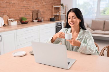 Mutfaktaki masada elinde bir fincan kahve ve dizüstü bilgisayarla genç bir kadın.
