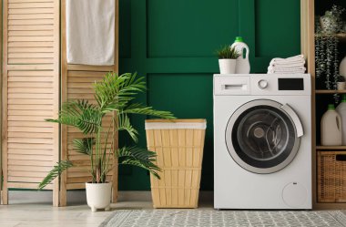 Çamaşır makinesi, katlanan ekran ve yeşil duvarın yanındaki çamaşır sepeti.