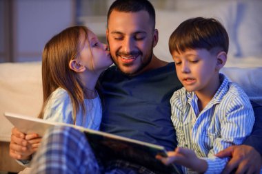 Baba ve küçük çocukları yatak odasında masal okuyorlar.