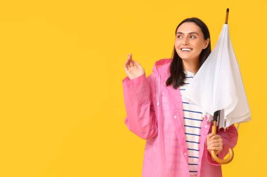 Yağmurluk giymiş, sarı arka planda şemsiyesi olan mutlu genç bir kadın.