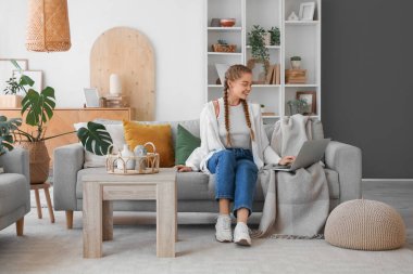 Genç bir kadın oturma odasındaki gri koltukta dizüstü bilgisayar kullanıyor.