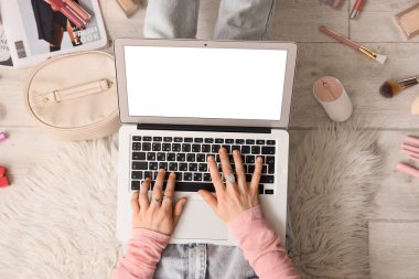 Hafif ahşap zeminde dizüstü bilgisayar ve makyaj ürünleri kullanan kadın güzellik blogcusu, üst görünüm