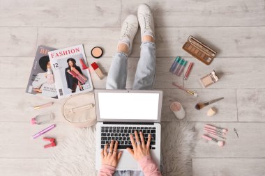 Hafif ahşap zeminde dizüstü bilgisayar ve makyaj ürünleri kullanan kadın güzellik blogcusu, üst görünüm