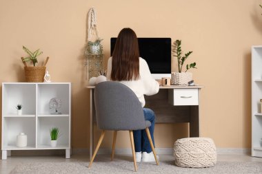 Bej duvarın yanındaki masada modern bilgisayarla çalışan genç bir kadın.