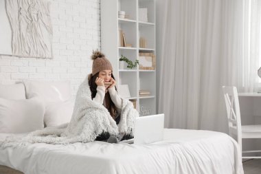 Sıcak ekoseli ve laptoplu donmuş genç kadın yatak odasında oturuyor.