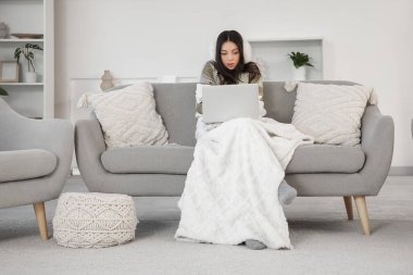 Evdeki kanepede dizüstü bilgisayar kullanan, sıcak ekoseli dondurulmuş genç bir kadın.