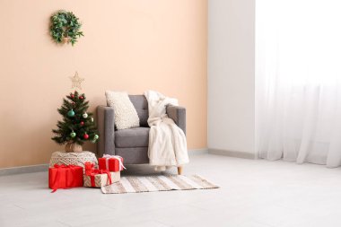 Koltuk ve küçük Noel ağacı. Odada bej duvarın yanında hediyeler var.