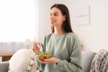 Sebze salatası ve çatallı genç bir kadın şık oturma odasında kanepede oturuyor.