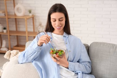 Elinde bir kase sebze salatası ve çatalla oturma odasında kanepede oturan genç bir kadın.