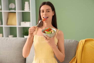 Oturma odasındaki kanepede sebze salatası yiyen genç bir kadın.