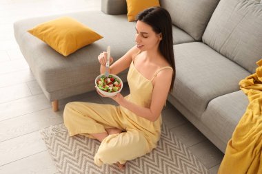 Genç bir kadın oturma odasında yerde sebze salatası yiyor.