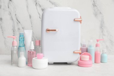 Küçük kozmetik buzdolabı ve kozmetik ürünleri mermer duvarın yanındaki masada.
