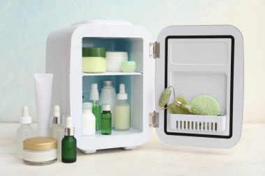 Açık mavi arka planda kozmetik ürünleri olan küçük bir kozmetik buzdolabı