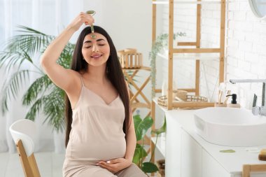 Genç hamile kadın banyoda yüz masajı aleti kullanıyor.