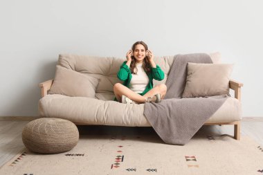 Kulaklıklı güzel bir kadın evdeki gri kanepede müzik dinliyor.