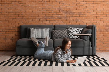 Genç ve güzel bir kadın evdeki siyah kanepenin yanında dizüstü bilgisayar kullanıyor.