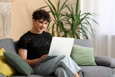 Genç transseksüel kadın evdeki kanepede dizüstü bilgisayar kullanıyor.