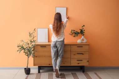Evdeki turuncu duvara boş bir çerçeve asan genç bir kadın.