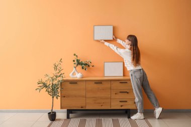 Evdeki turuncu duvara boş bir çerçeve asan genç bir kadın.