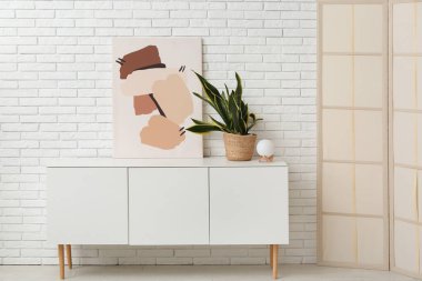 Beyaz tuğla duvarda asılı resim, çekmeceler, katlanır ekran ve oda çiçekleri.