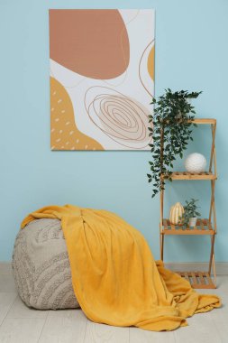 Mavi duvarda asılı resim, ekoseli puf, raf ünitesi ve oda bitkisi.