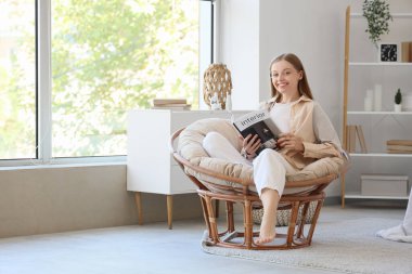 Evdeki koltukta iç tasarım dergisi okuyan genç bir kadın.