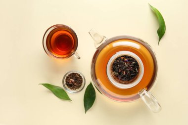 Şık çay demliği, bir fincan çay ve bej arka planda yeşil yapraklar.