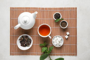 Çay demliği, çay fincanı, bambu hasırı ve açık arkaplanda yeşil yapraklarla güzel bir kompozisyon.