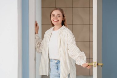 Güzel genç bir kadın evinin açık kapısında duruyor.