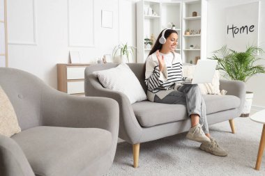 Kulaklıklı, dizüstü bilgisayarlı, evdeki kanepede sohbet eden güzel bir genç kadın.