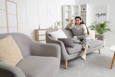 Kulaklıklı, dizüstü bilgisayarlı, evdeki kanepede sohbet eden güzel bir genç kadın.