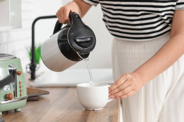 Güzel genç bir kadın modern elektrikli çaydanlıktan sıcak suyu mutfaktaki bardağa dolduruyor.