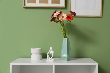 Vazodaki gerberalar ve heykelcikler yeşil duvarın yanındaki raflarda.