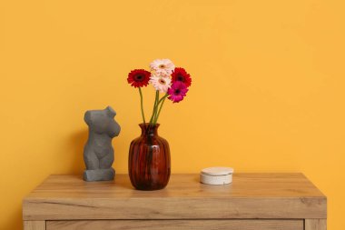 İçinde Gerbera çiçekleri olan vazo ve odadaki sarı duvarın yanındaki çekmecelerin üzerindeki kadın figürü. Yakın çekim.