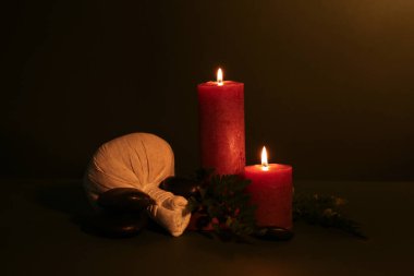 Masaj çantalı kompozisyon, yanan mumlar, spa taşları ve koyu renk arka planda eğreltiotu yaprakları.