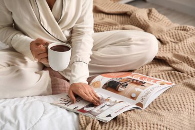 Evdeki yatakta bir fincan sıcak çikolata okuma dergisiyle güzel bir genç kadın, yakın plan.