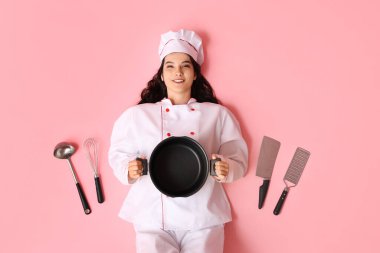 Farklı mutfak aletleri ve pembe arka planda duran çömleği olan mutlu genç bayan aşçı.