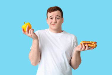 Mavi arka planda dolmalık biberli ve sosisli sandviç yiyen düşünceli genç bir adam. Kilo alma kavramı